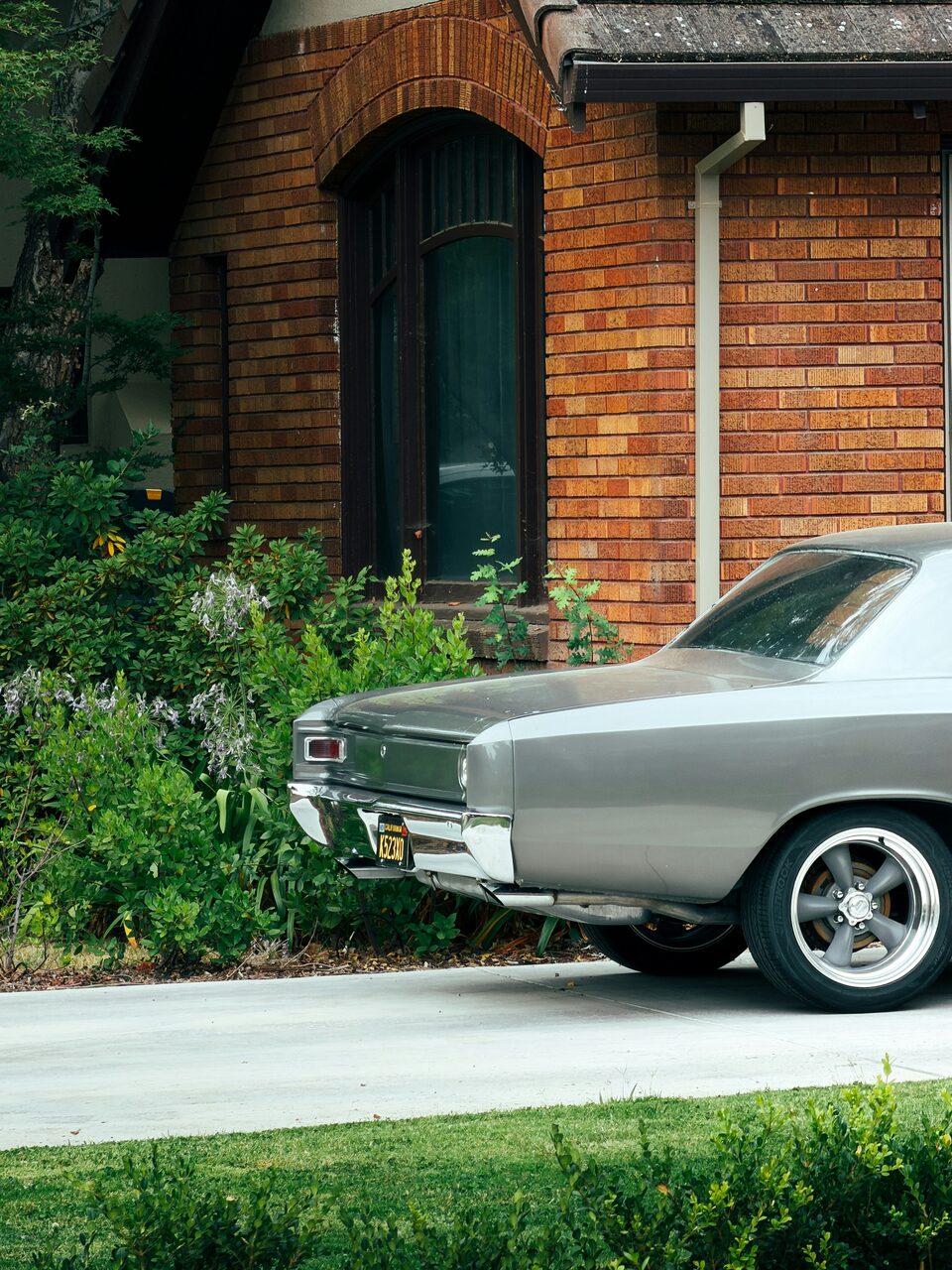 A silver classic car beside a brick house and garden